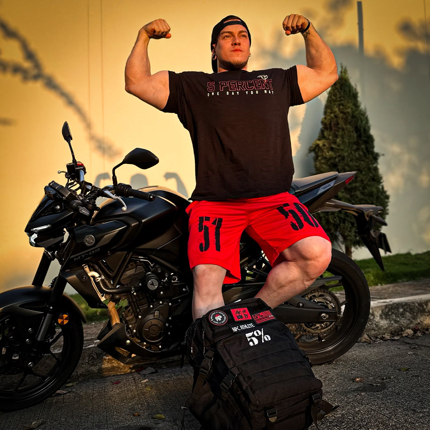 Muscular man flexing arms in 5% Nutrition apparel beside black motorcycle at sunset