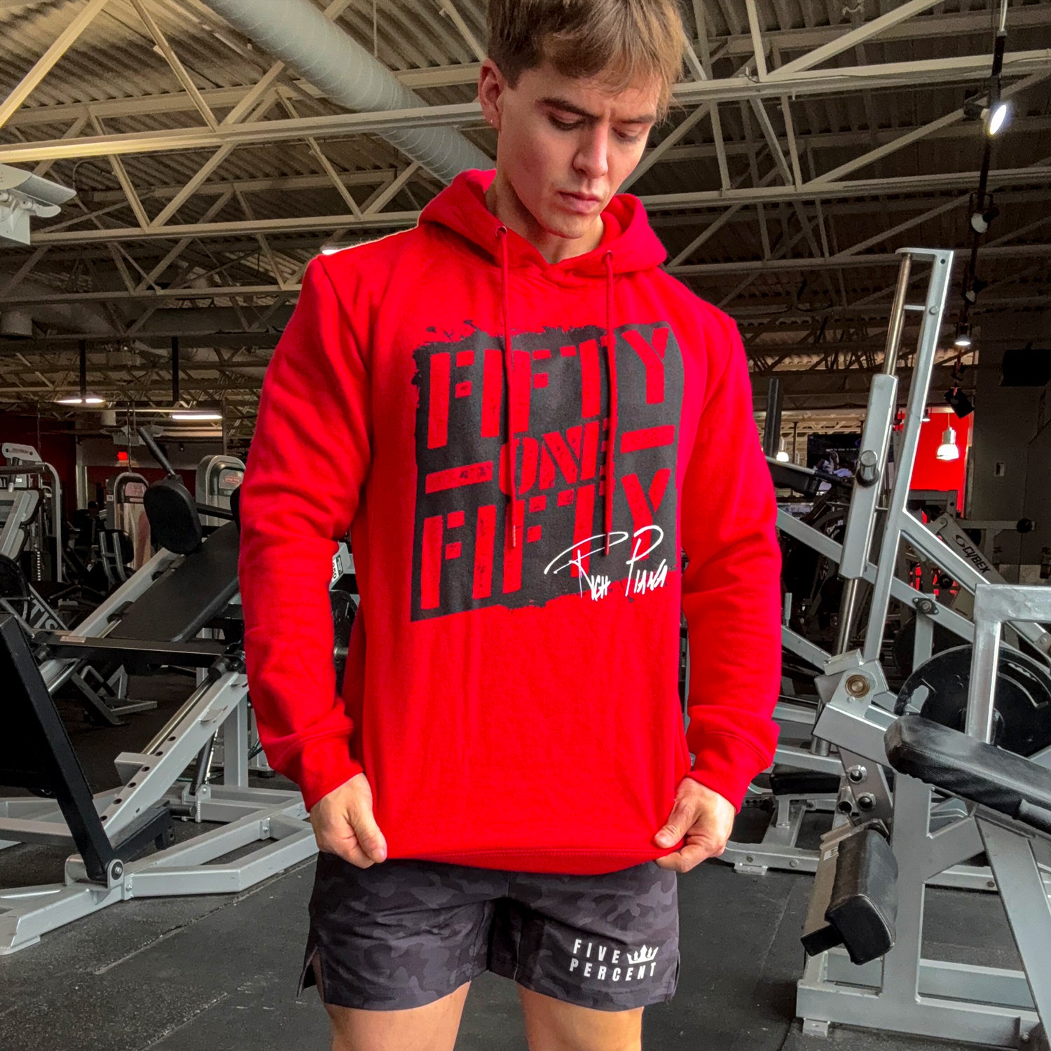 Red hoodie with 'FIFTY ONE FIFTY' graphic and Rich Piana signature, worn in gym setting with workout shorts.