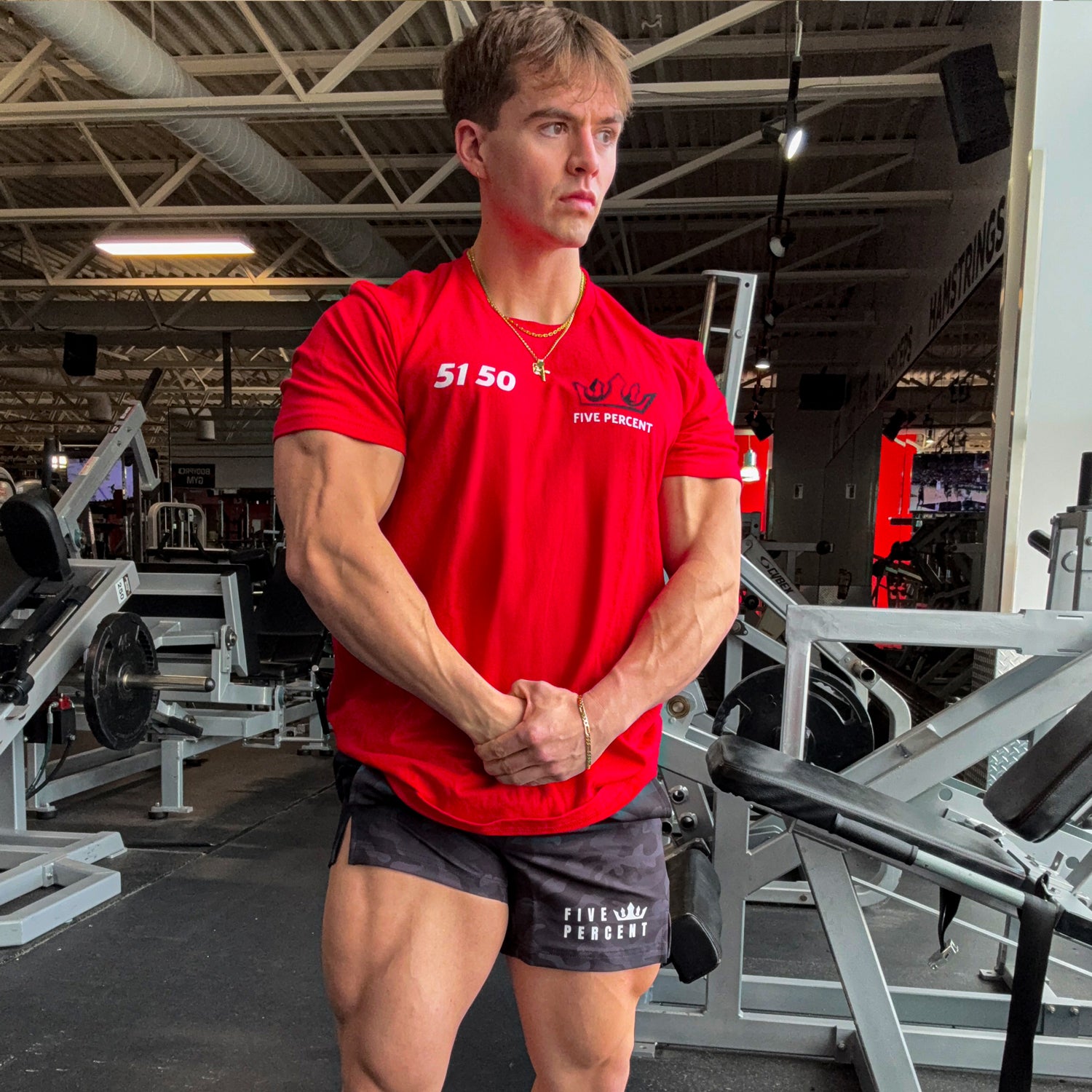 Muscular man in red Five Percent gym shirt and camo shorts, posing in fitness center with equipment
