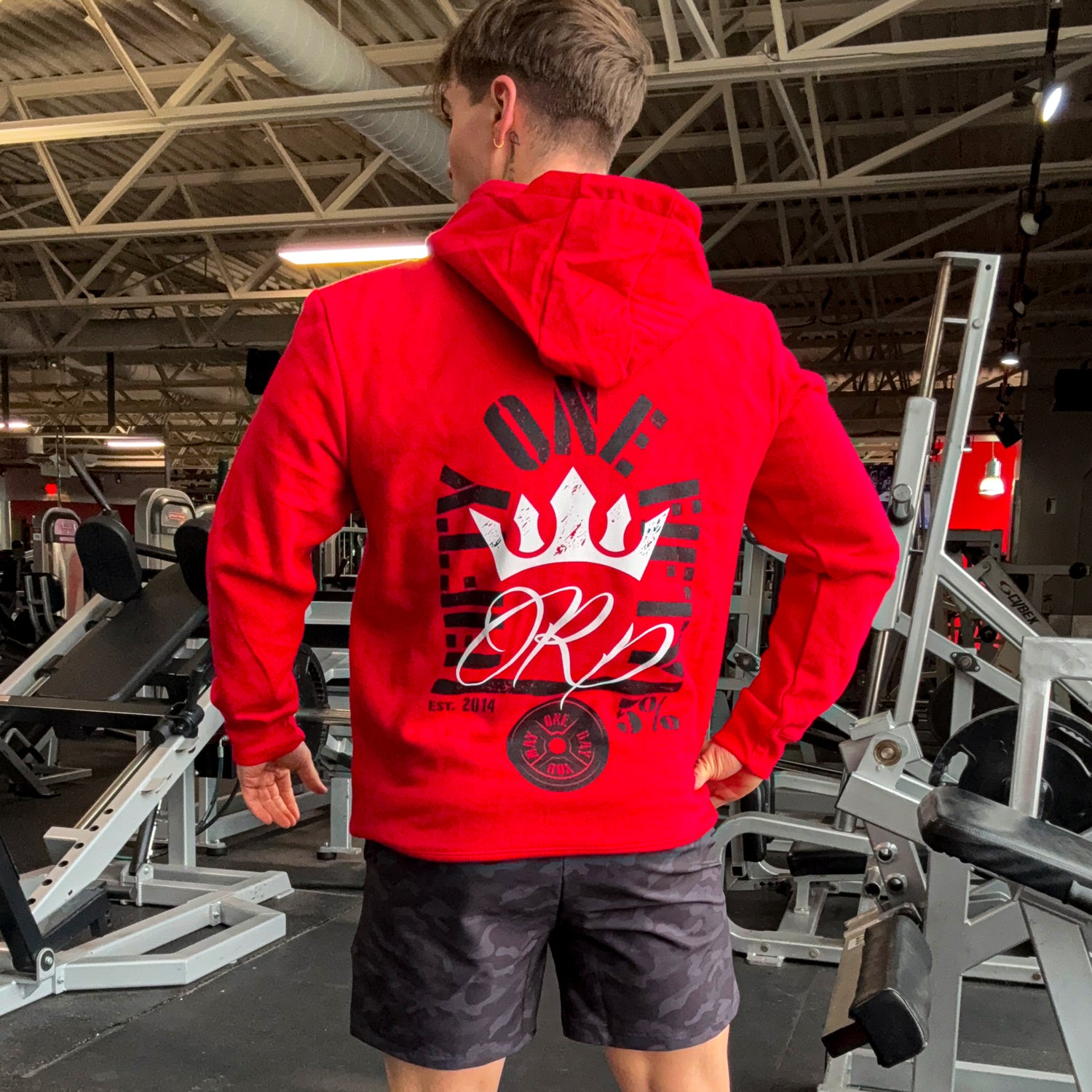 Man in red gym hoodie with crown graphic, 'One Life One Rep' text, and 5% logo, standing in fitness center